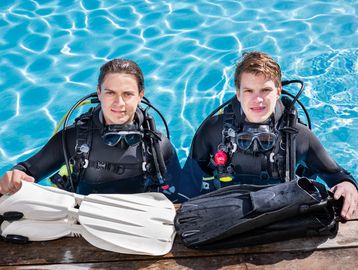 Two scuba divers holding fins at the pool edge, smiling.