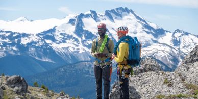 Two climbers standing on rocky terrain with snowy mountains in the background.