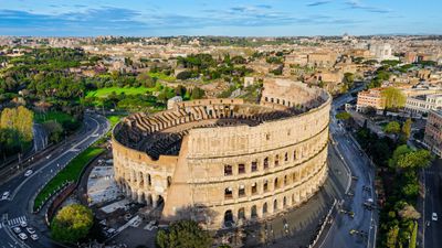 Aerial view of the Colosseum in Rome surrounded by cityscape and greenery.
