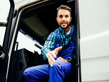 A smiling truck driver reaching out for a handshake from his truck cabin.