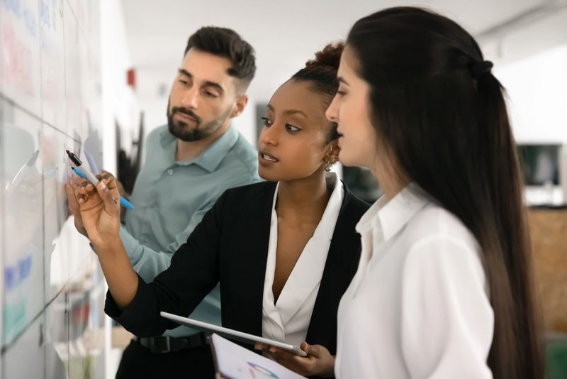 Diverse workmates working together on business plan, writes on whiteboard standing in modern office, engaged in visual representation of data or market trends analysis, reviewing key insights on board