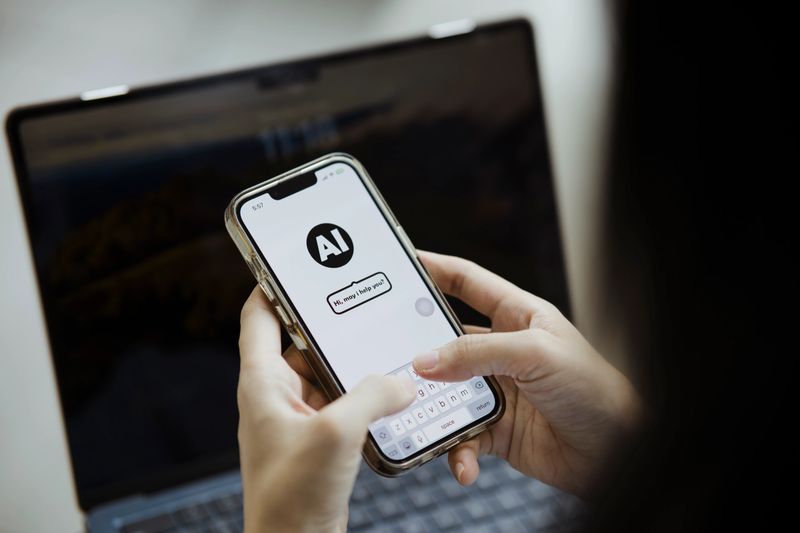 A person interacts with an AI chatbot on a smartphone. The screen displays a conversation interface with the text "Hi, may I help you?" suggesting the use of an AI assistant app. A laptop is visible in the background, emphasizing a tech-savvy environment.