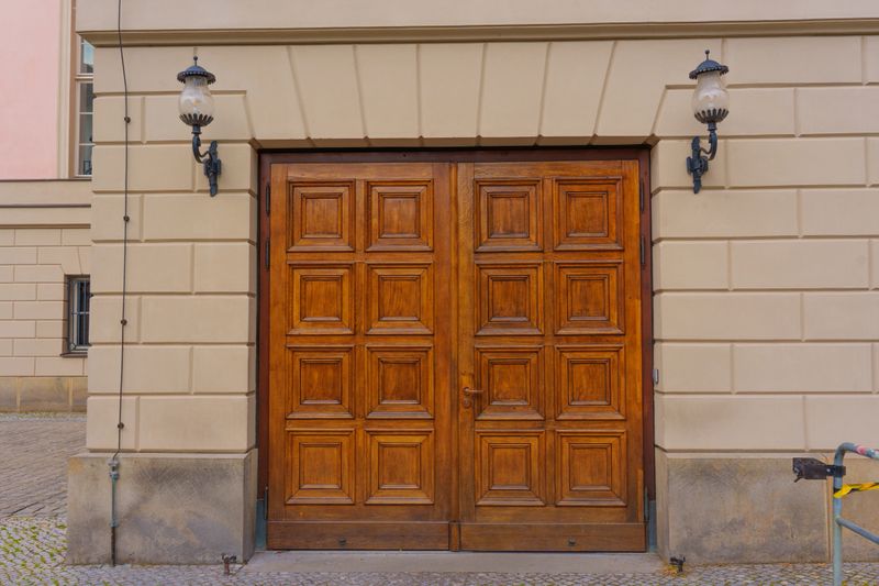 Pair of ornate wooden doors featuring detailed paneling, flanked by vintage lanterns, set against a light-colored stone wall, showcasing historic architectural elements.