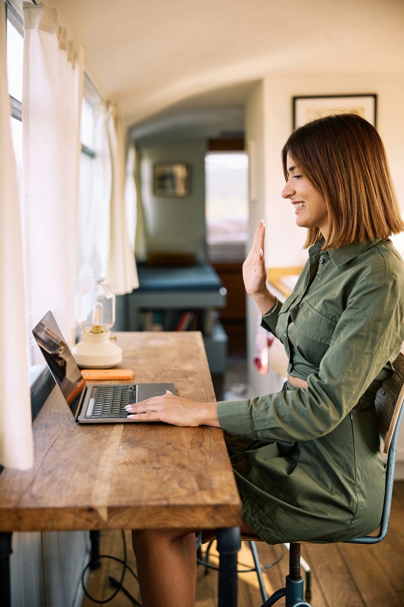 A cheerful woman in a green dress works on her laptop in a well-lit, cozy vacation home with natural light streaming through large windows and waving at the laptop during conference call