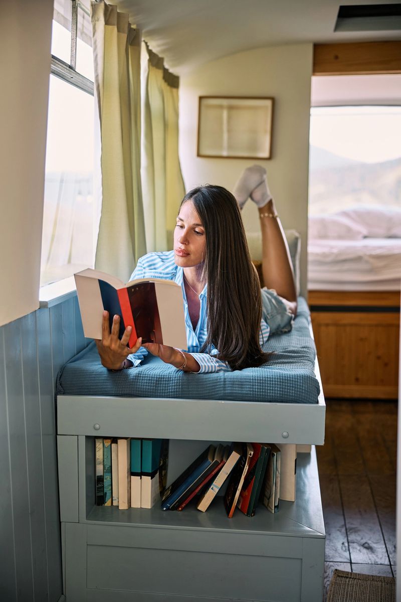 A young woman with long dark hair is lying on a camper van bed, surrounded by books, reading in a peaceful and cozy interior.