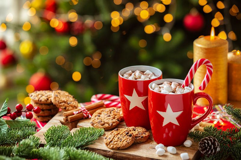 Hot chocolate marshmallow mug and chocolate chips cookies on Christmas decorated table near illuminated Christmas tree. Sweet food for December Holidays.