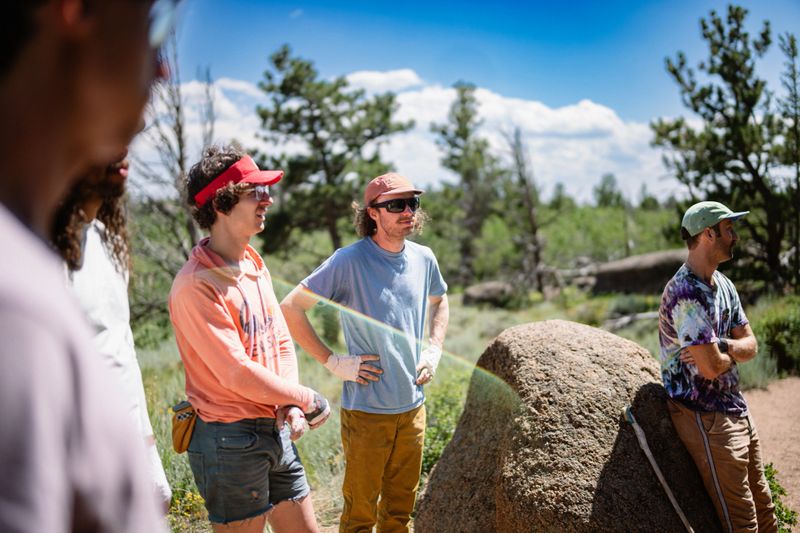 A group of male friends chat cheerfully as they watch their friend rock climb at Vedauwoo Climbing Area in Wyoming during a weekend camping trip for a bachelor party.