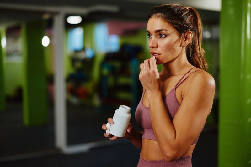 Young sportswoman taking dietary supplement pill after intensive workout in gym
