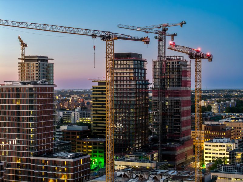 Evening aerial view of the Zuidas business district in Amsterdam South, where multiple high-rise towers are under construction. Tower cranes rise above illuminated glass facades as city lights shimmer against a fading sky, highlighting rapid urban growth, real-estate investment, and the Netherlands’ modern architectural ambitions.
