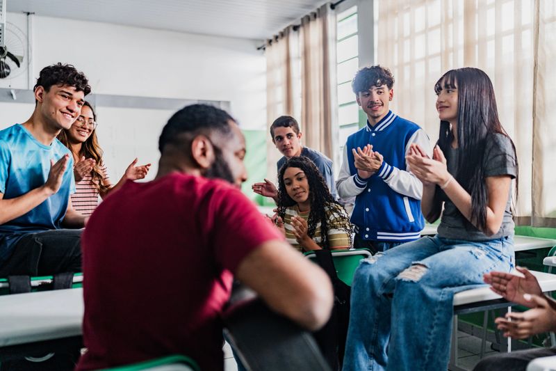 Inside a school classroom, a teacher plays the guitar while a group of young students joins in by clapping and listening attentively. The musical activity takes place in a relaxed and participative setting, with students sitting in a circle around the teacher. The scene conveys engagement, collaborative learning, and an encouraging artistic atmosphere within the educational environment