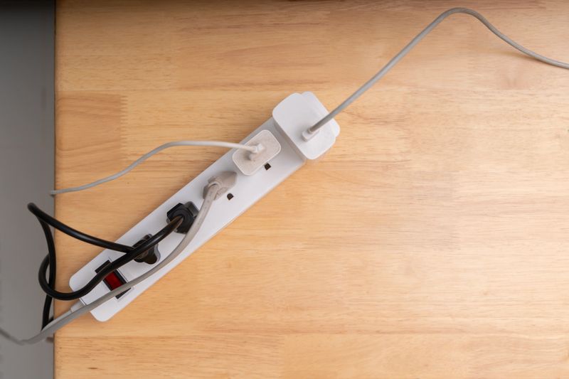 Top view of white power strip with several plugs and tangled black and white cables on wooden table. Concept of electricity, energy usage, power connection, home office, and modern devices.