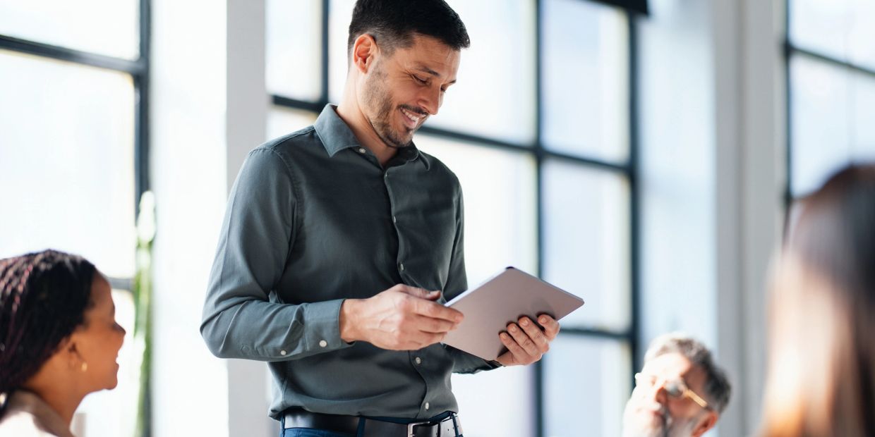Man smiling while using a tablet in a bright office meeting.