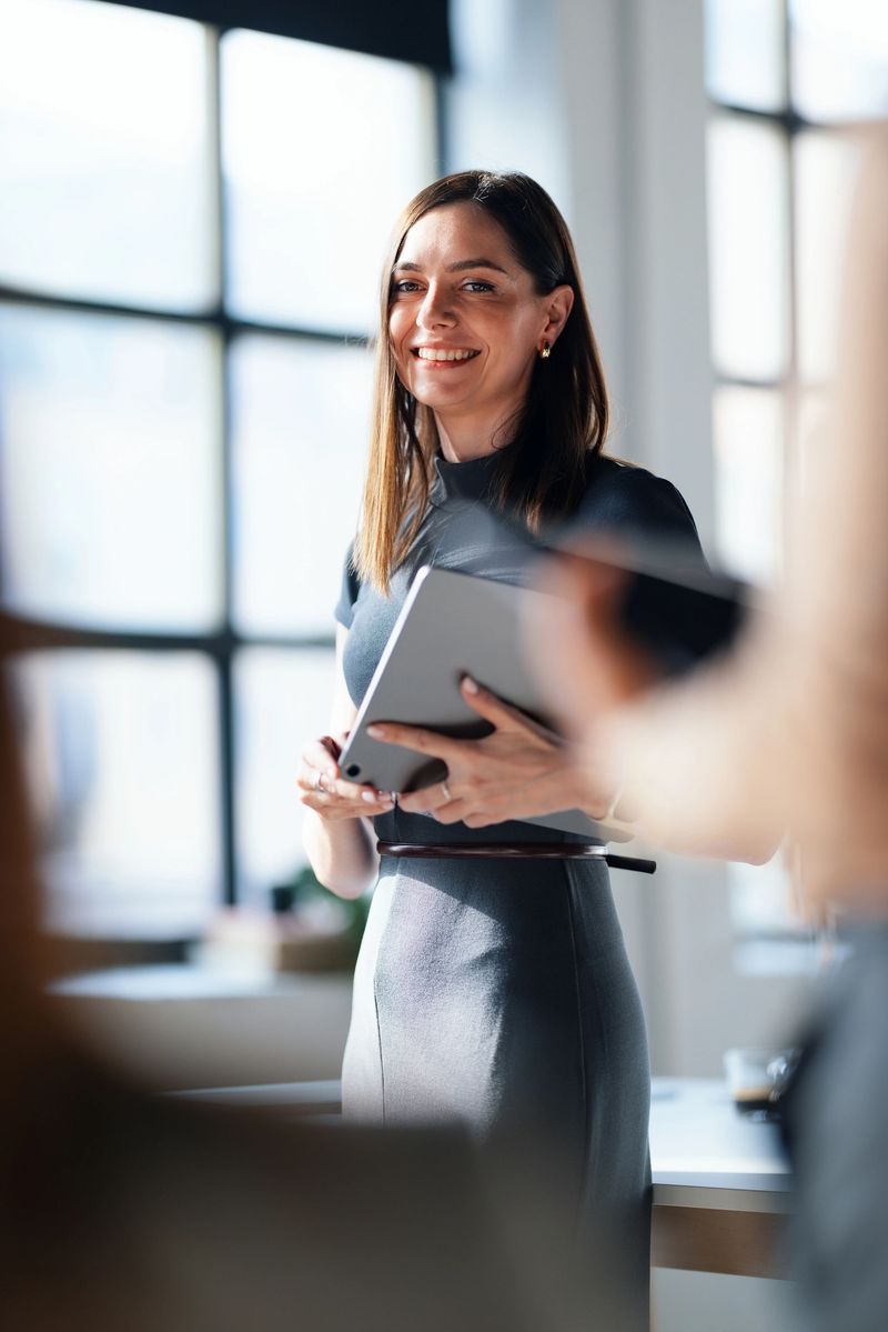 A professional woman smiling confidently while holding a tablet in a bright office. The sunlit workplace and her demeanor portray a sense of motivation, ambition, and teamwork.