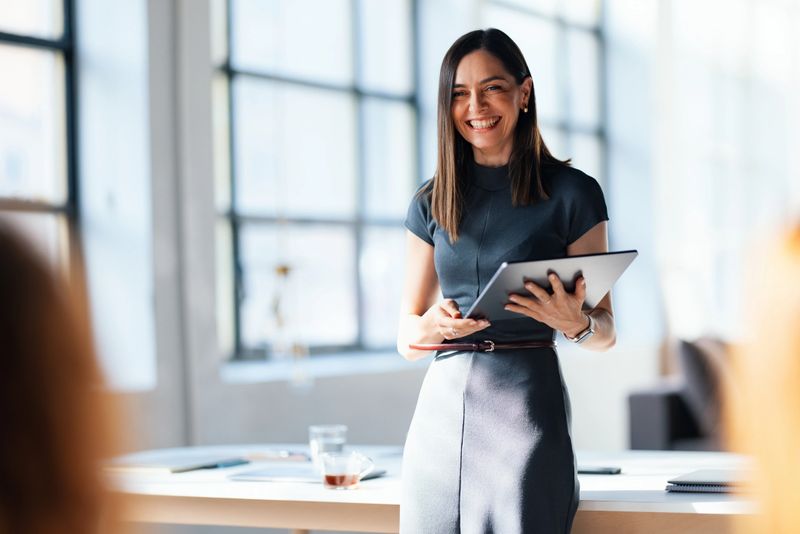 Smiling professional woman presenting in an office environment, holding a tablet, showcasing confidence and expertise. The natural lighting and supportive atmosphere highlight teamwork and career-oriented aspirations.