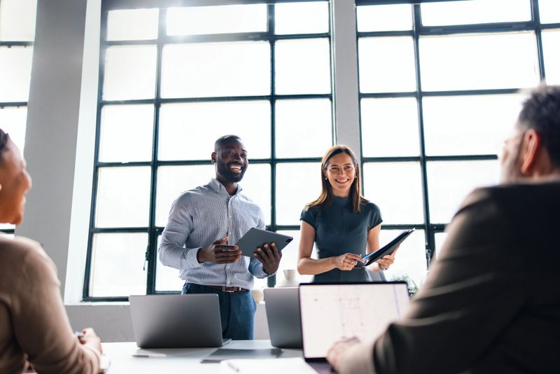 A diverse group of professionals collaborating in a brightly lit office during a business meeting. Smiling individuals, laptops, and teamwork represent a productive and inclusive workplace culture.