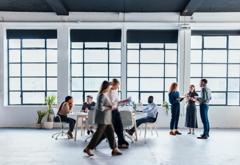 An open office environment showcasing professionals engaging in discussions and teamwork activities, surrounded by creative decor with large windows letting in natural light.
