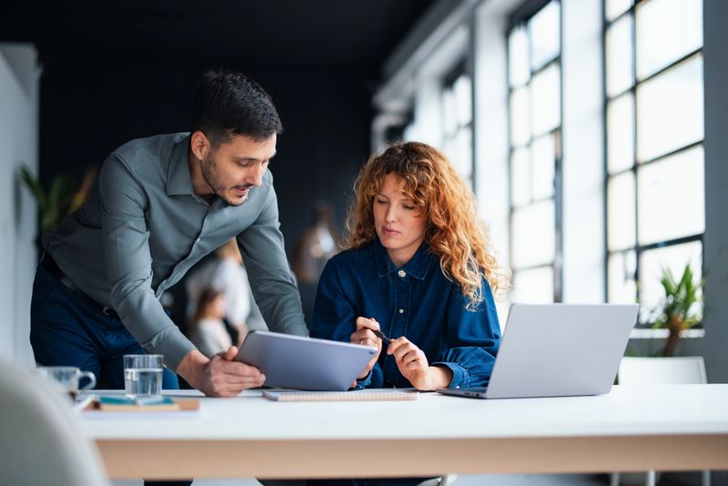 Two colleagues collaborate on a project in a stylish office with natural lighting and spacious workspace, emphasizing teamwork, creativity, and productivity.