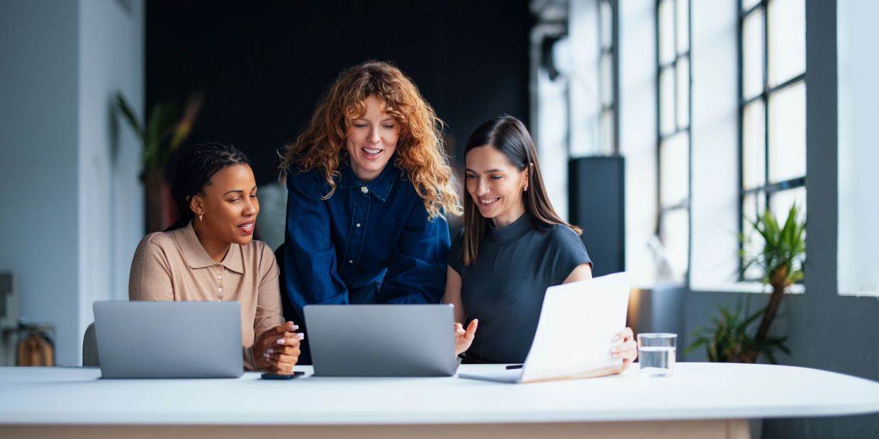 Three women collaborating in an office with laptops.