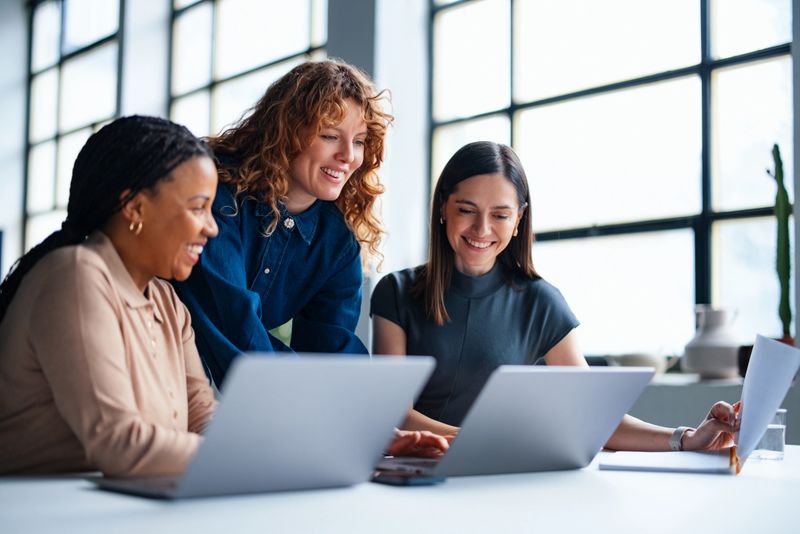 Three women are gathered in a bright office space, collaborating and discussing ideas while engaging with laptop computers. This setting conveys teamwork, efficiency, and collaboration within a professional business environment.