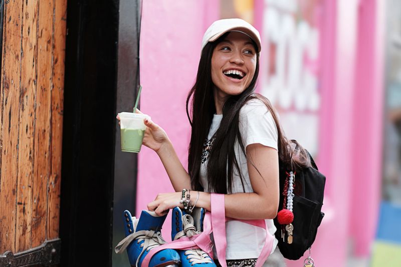 Mixed-race woman with a relaxed, streetwear vibe enjoys a cool iced matcha latte while returning home from a skate session. Wearing a baseball cap and casual t-shirt, she embodies effortless urban style and Gen Z skater culture. Her joyful expression and carefree energy capture modern youth lifestyle, wellness trends, and the spirit of independence. Perfect for themes like female skaters, iced drinks, street style, city living, and active lifestyle content.