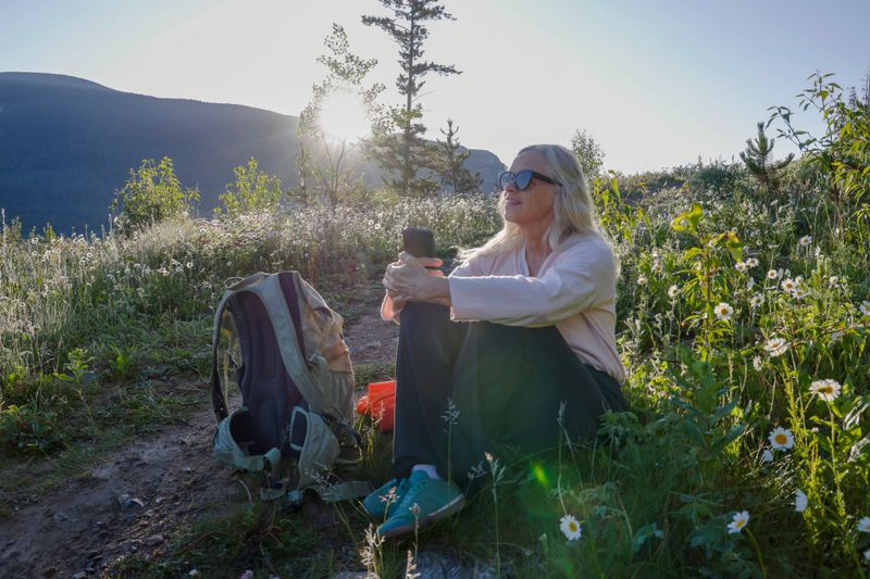 Mature woman relaxes on side of trail in mountain meadow at sunrise, she looks off to distant scene, Canadian Rocky Mountains