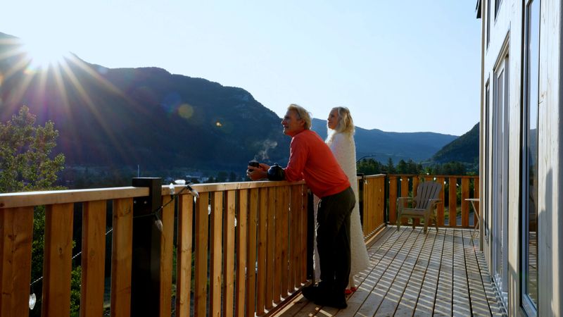 Distant view of mature man and woman, wrapped in blanket, enjoy a cup of hot tea, on cabin balcony at sunrise, Crowsnest Pass, Alberta