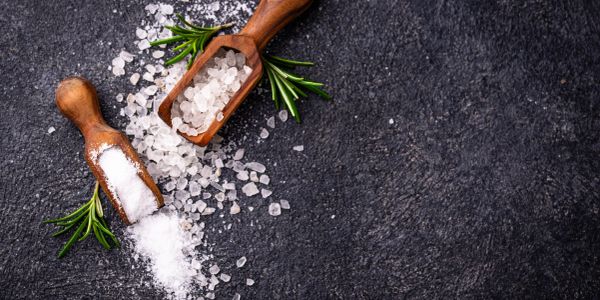 Wooden scoops with coarse and fine salt on dark surface with rosemary.