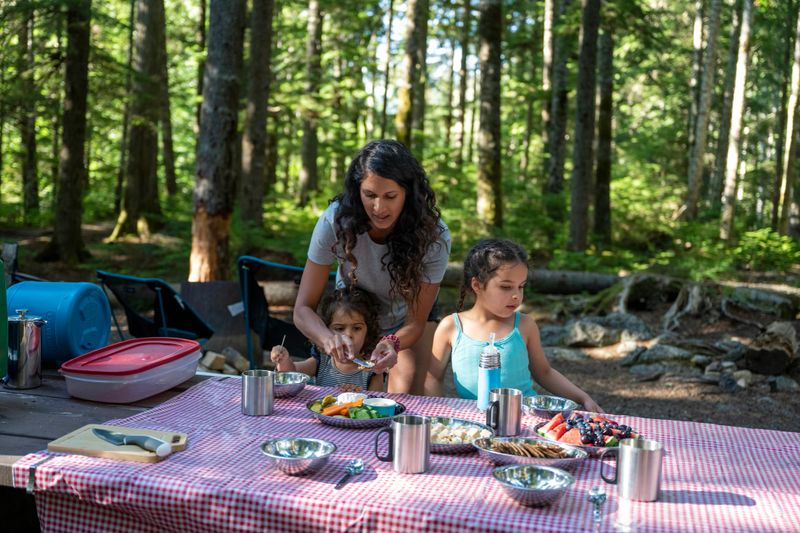 Mother and young daughters eat at campsite picnic table in forest, British Columbia