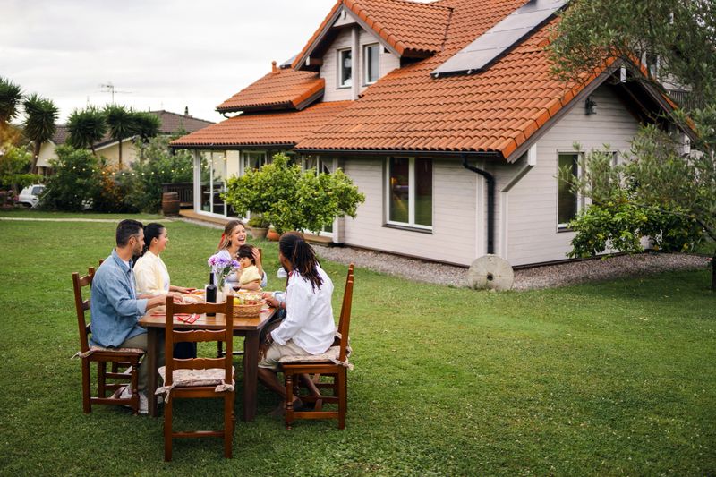 Multi-ethnic family is gathering around a table in their backyard, enjoying a meal together in front of their house with solar panels