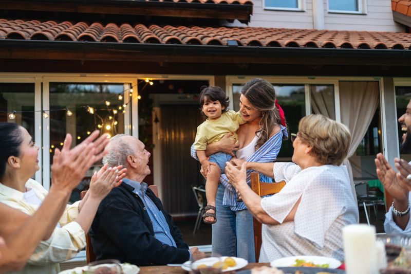 Large multi-generational family gathering, rejoicing newborn arrival with cheerful conversations, hugging relatives, enjoying sunset garden ambiance