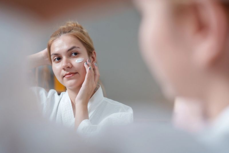 Young woman applies skincare cream to her face while looking mirror, showcasing moment of self care and beauty. This routine promotes skin health and helps brighten her complexion, enhancing her