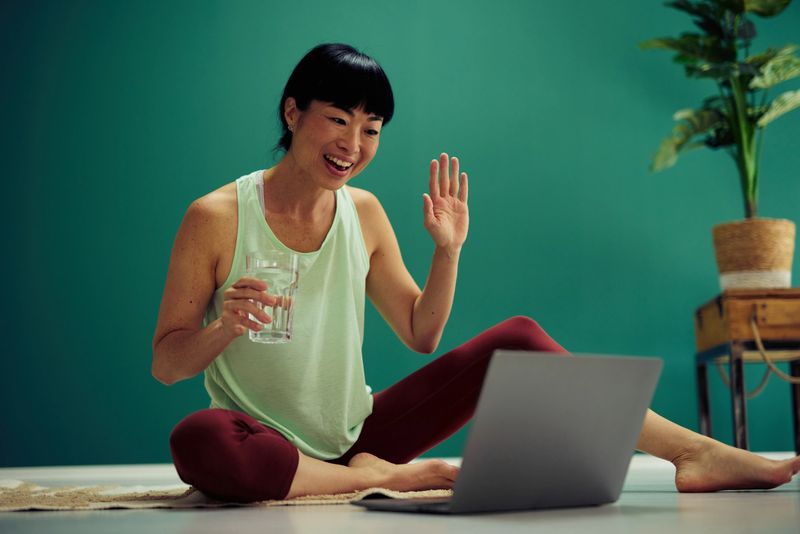 Happy mid adult woman practicing yoga at home following online class on laptop, she is smiling and performing a low lunge pose on a rug in front of a green wall