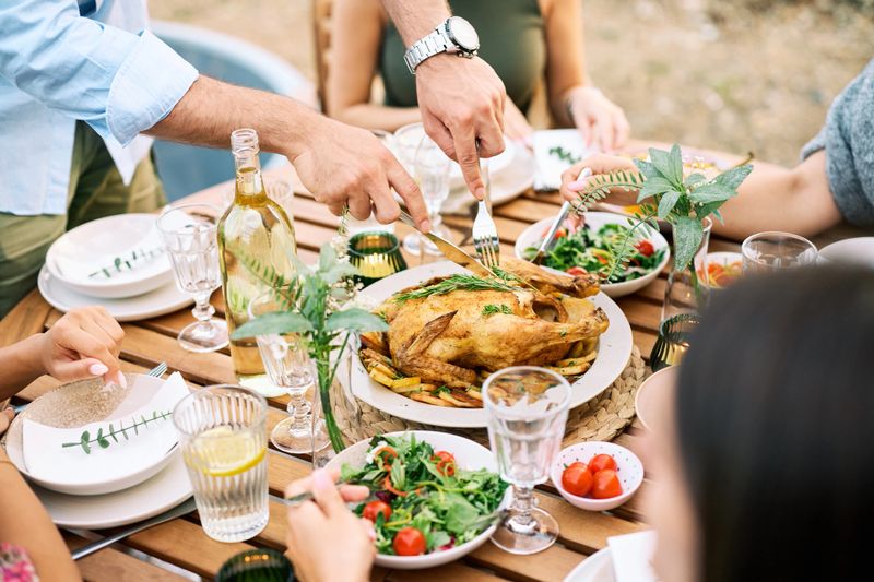 A group of friends enjoys a shared meal outdoors in a serene natural setting. The table is adorned with fresh salads, a roasted chicken, and wine, creating a warm and inviting atmosphere of togetherness.