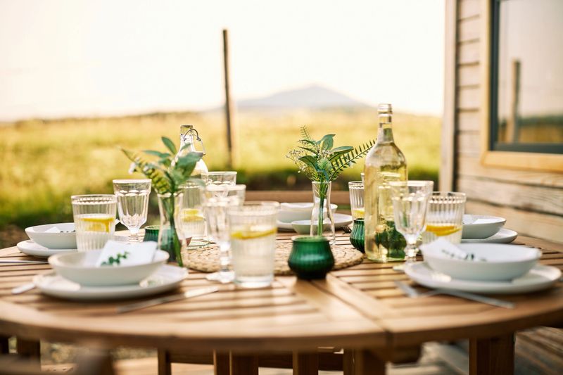 An inviting outdoor dining table with elegant glassware, white dishes, greenery, and a bottle of wine set against a bright, sunny backdrop.