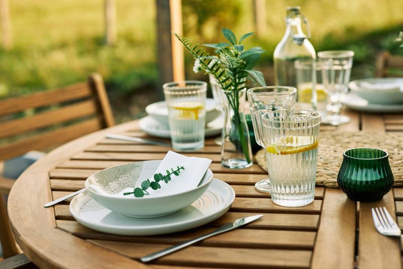 A cozy outdoor wooden table set for lunch, featuring fresh lemon water, plates, and greenery accents under the warm daylight in nature.