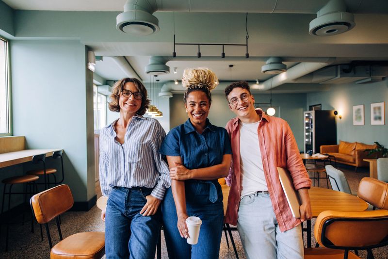 A group of three coworkers of diverse backgrounds are smiling together in a coworking office. Their cheerful dispositions reflect positivity and camaraderie.