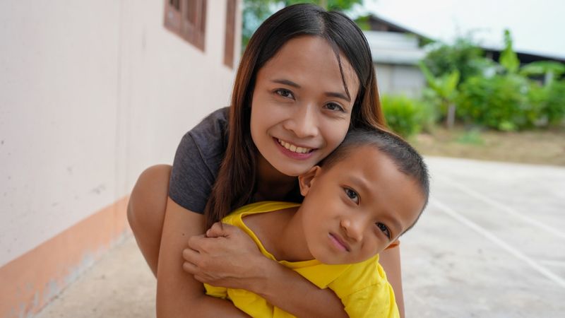 A joyful woman embraces young boy outdoors, showcasing warm and loving bond. background features greenery and simple building