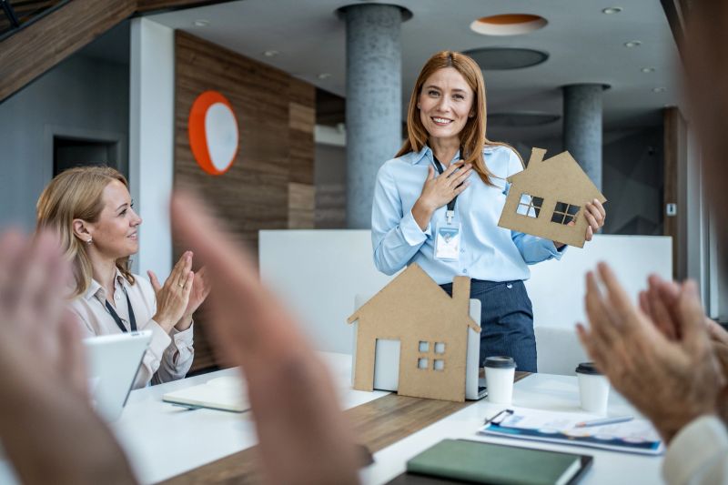 Businesswoman is holding a cardboard cutout of a house while receiving applause from her colleagues after a successful presentation in a modern office
