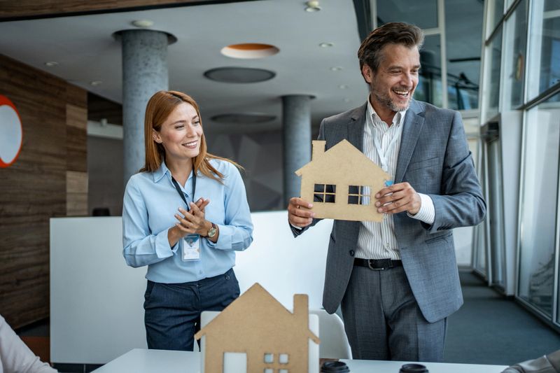 Real estate agent holding a cardboard house model, presenting it to a potential buyer in a modern office, discussing property investment and homeownership