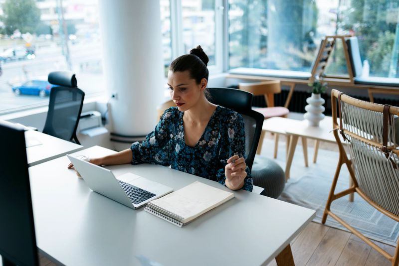 Young adult businesswoman concentrating on her laptop, taking notes in a notebook, in a bright, modern office space