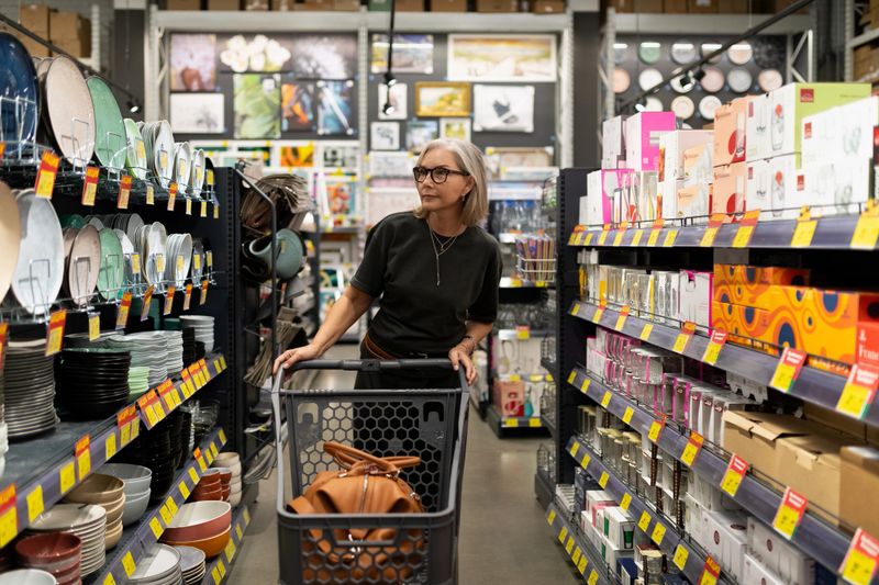 A stylish mature woman explores a contemporary home goods store, selecting dinnerware, pots, decor items, and houseplants for her living space while pushing a shopping cart.