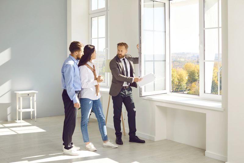Handsome bearded male real estate agent in suit with documents in his hands advises young couple before buying new house, demonstrates view from the window. Three people are standing in modern sunny apartment.