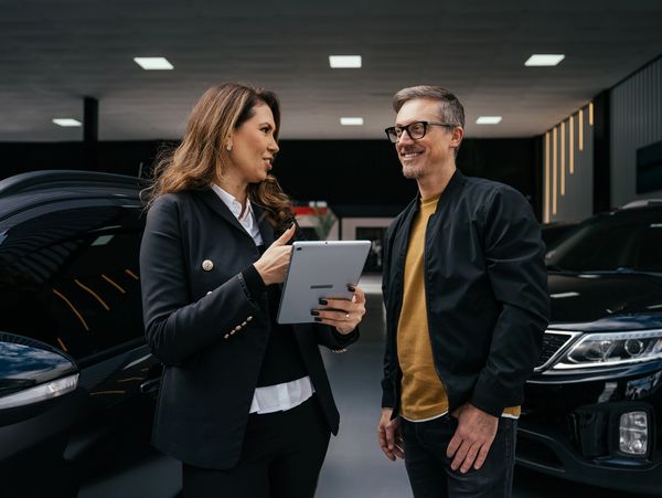 A woman shows a tablet to a man in a car showroom.