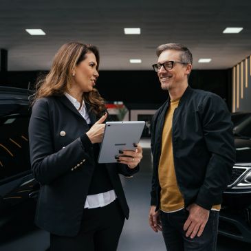 A woman shows a tablet to a man inside a car dealership.
