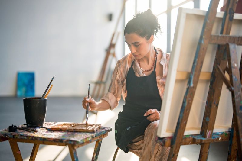 A young Middle Eastern female artist paints attentively in a bright studio. She wears casual clothing and an apron while working on a canvas, surrounded by art tools and natural light.