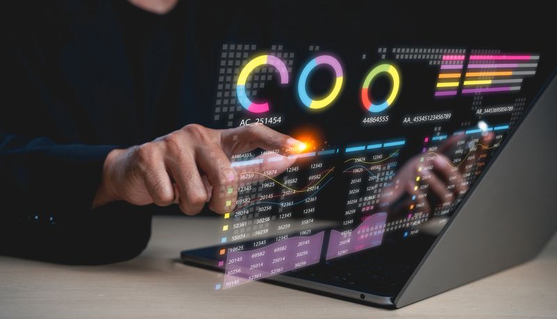 A person interacts with a futuristic transparent touchscreen displaying colorful charts and data analytics above a laptop.