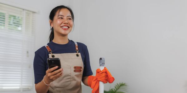 Smiling woman in apron holding phone and cleaning tool indoors.