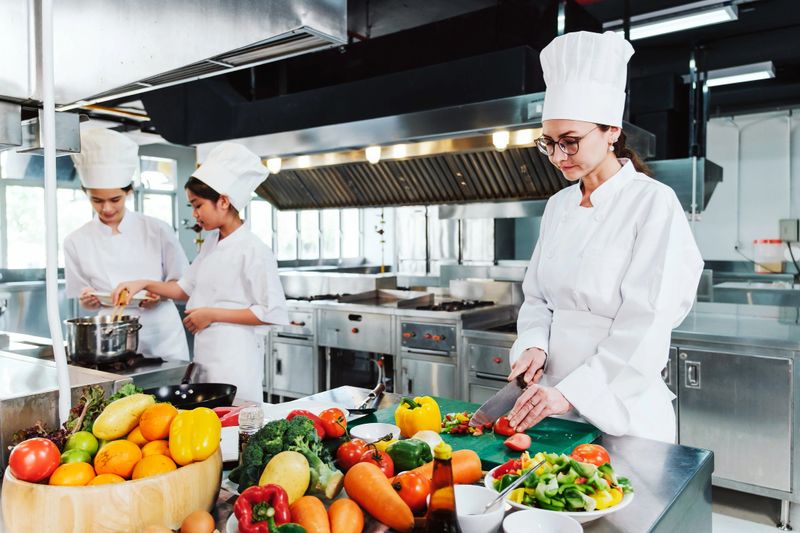 Chef in white uniform slicing fresh tomatoes on a green cutting board with chopped bell peppers in a professional kitchen, preparing ingredients for a colorful, healthy dish during cooking class.