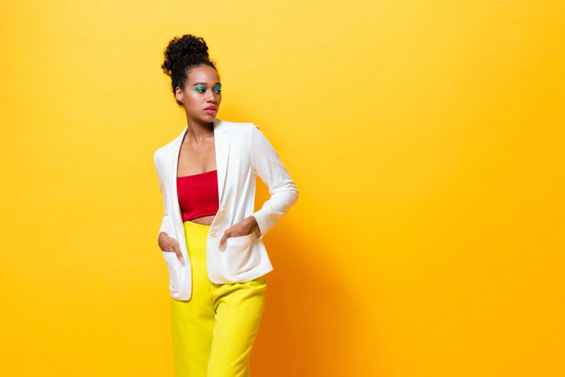 Stylish woman with bold make up in colorful outfit posing confidently against vibrant yellow background studio shot