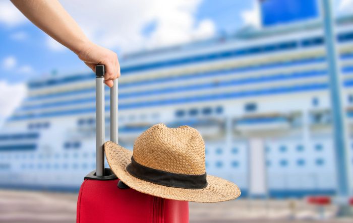 Hand holding luggage near a cruise ship with a straw hat.